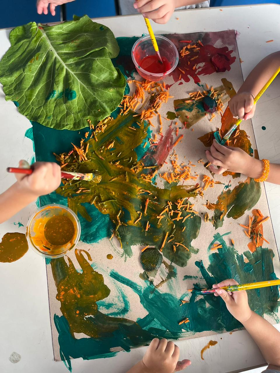 Crianças pintando uma tela com tinta, nas cores dos legumes que estão em cima da mesa